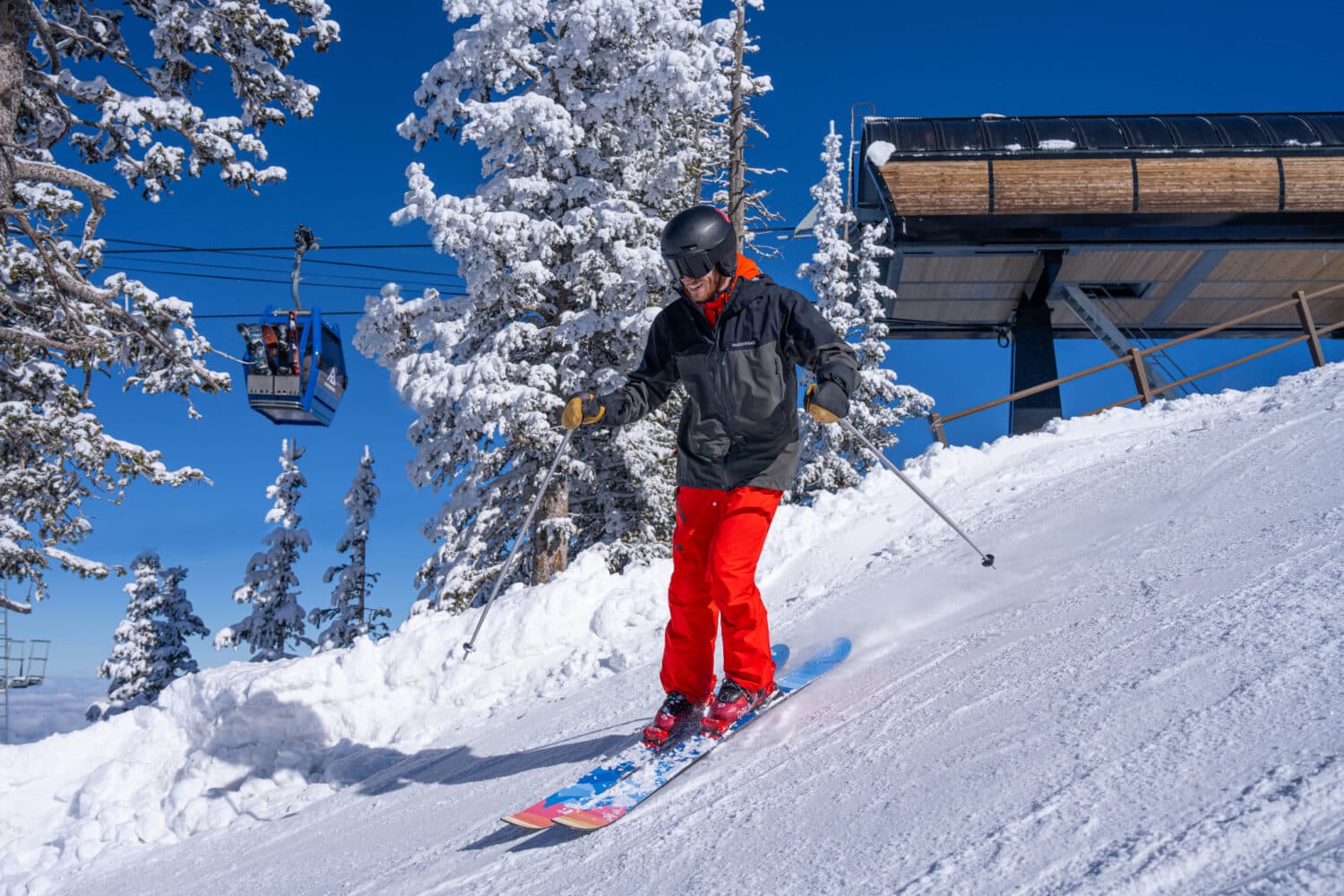 Skier smiling while riding down groomed run with Arizona Gondola passing in the background.